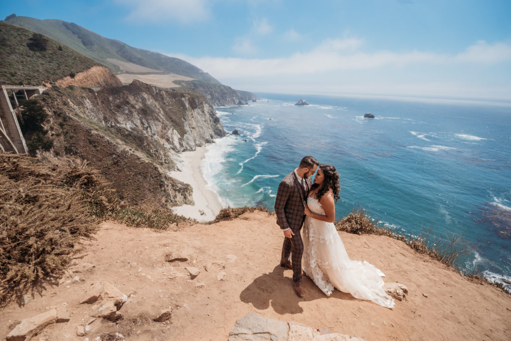 Couple snuggling on the cliffs of Big Sur with the waves and bridge behind them on a sunny day.